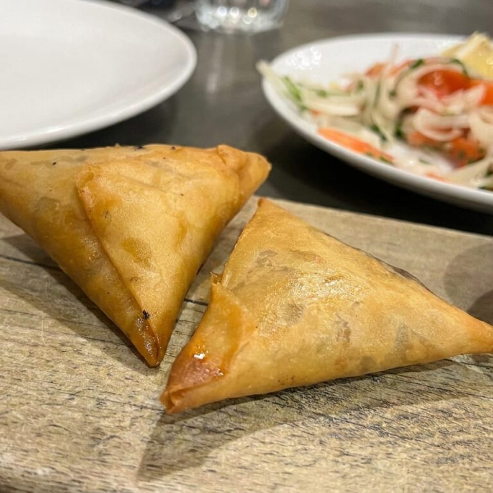 Two golden fried samosas sit on a wooden serving board with brown dipping sauce and a noodle salad in the background.