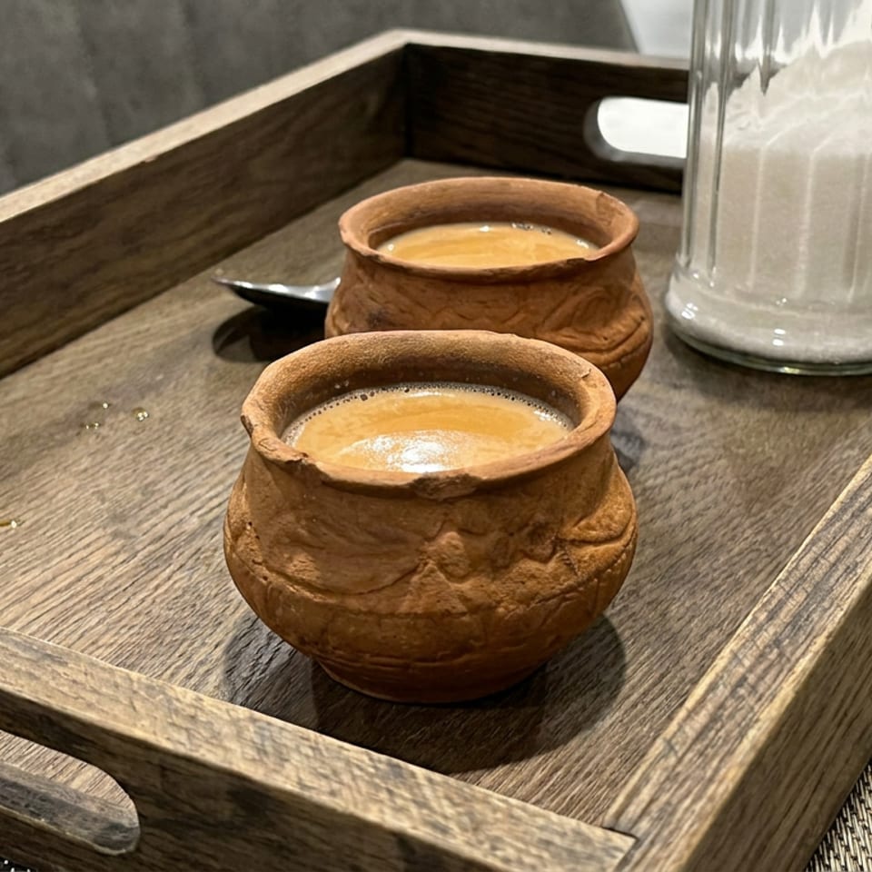Two rustic clay cups filled with frothy coffee sit on a wooden serving tray beside a glass sugar container.