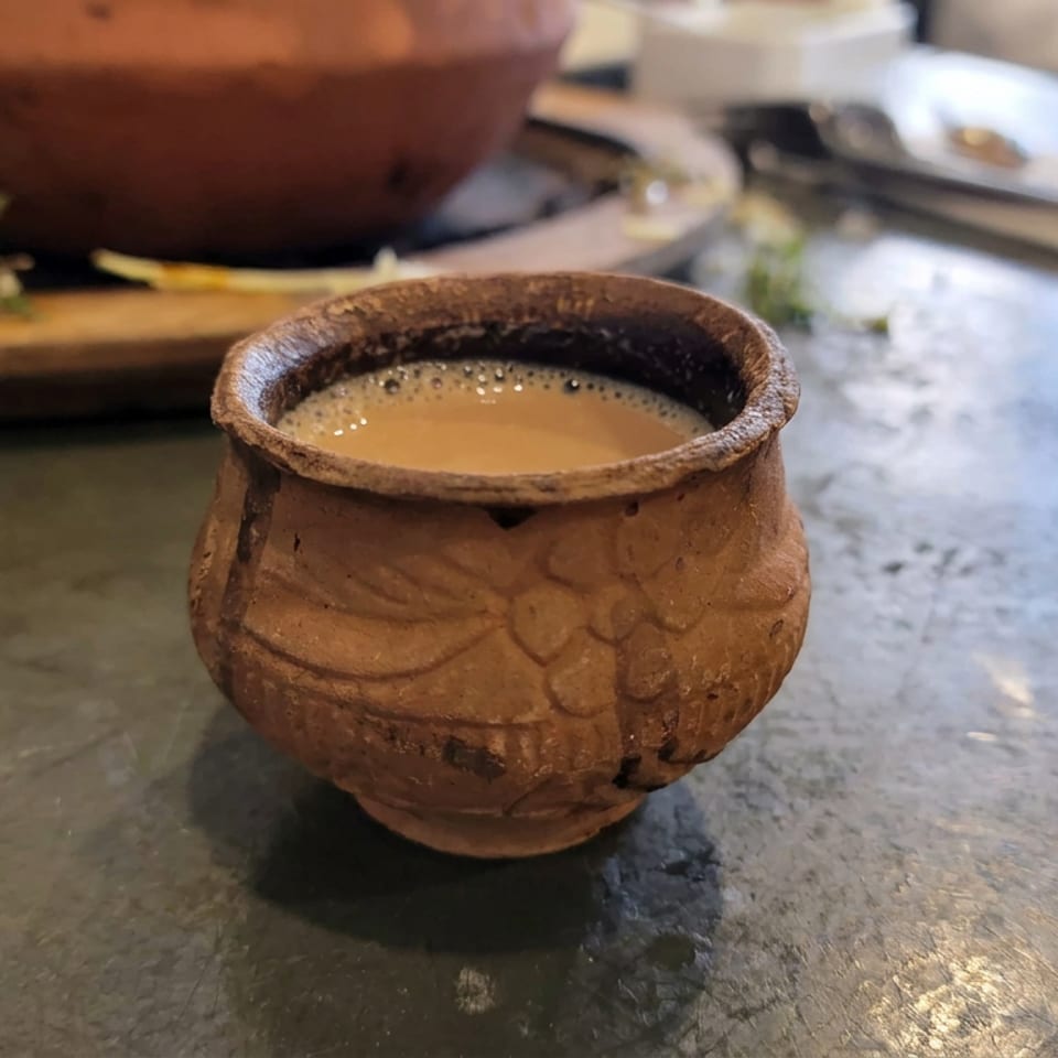 Rustic ceramic cup filled with frothy tea sits on a restaurant table, with a cutting board and herbs softly blurred behind it.