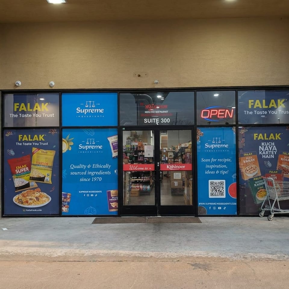 Falak storefront entrance with blue Supreme grocery advertisements, glass doors, brick columns, and shopping carts outside.